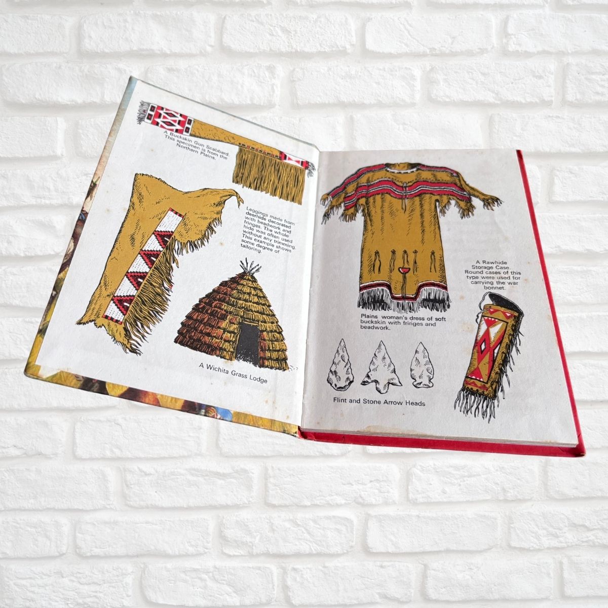 An open illustrated book or booklet laid flat against a white brick wall, showing drawings of Native American-style clothing, a lodge tent, and artifacts with small captions. No person or animal is present.