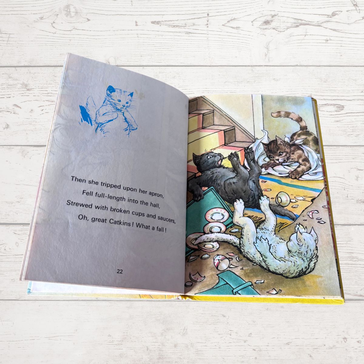 An open children's book lying on a light wood floor, showing a printed page with a blue sketch of a cat and a colorful illustration of cats playing in a room.