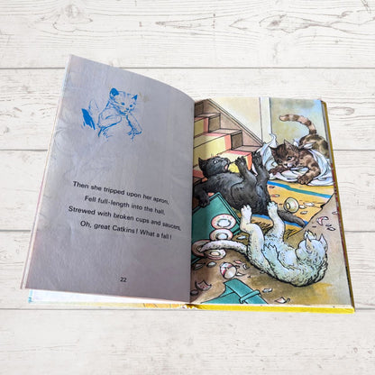 An open children's book lying on a light wood floor, showing a printed page with a blue sketch of a cat and a colorful illustration of cats playing in a room.