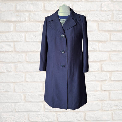 A navy blue knee-length coat with notched lapels and three front buttons, displayed on a mannequin torso against a white brick wall background.