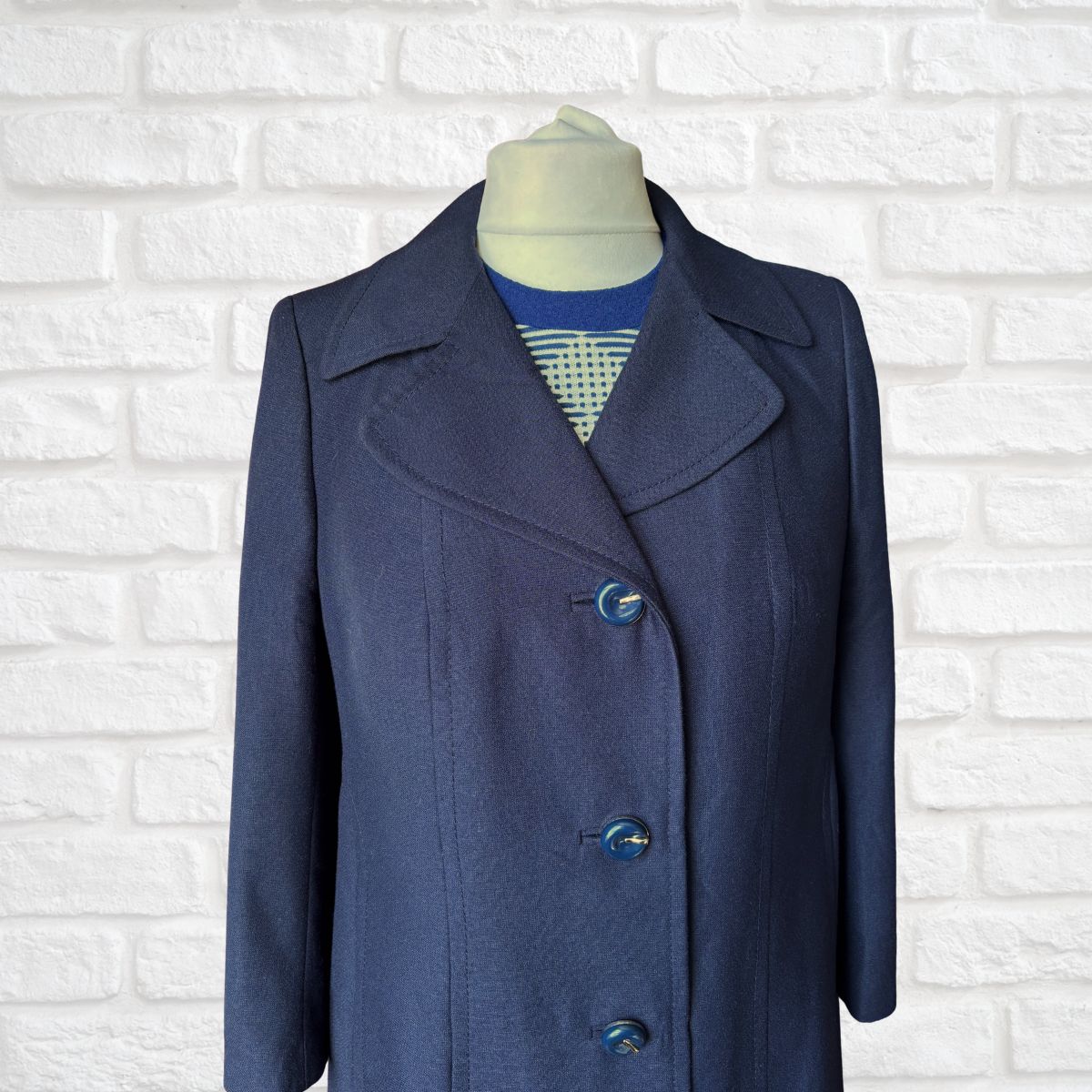 A navy blue single -breasted coat displayed on a mannequin torso against a white brick wall background.