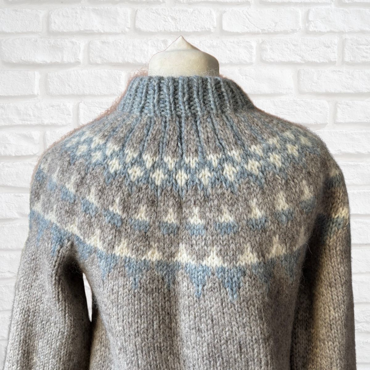 Knitted sweater with a pattern on a mannequin against a white brick wall.