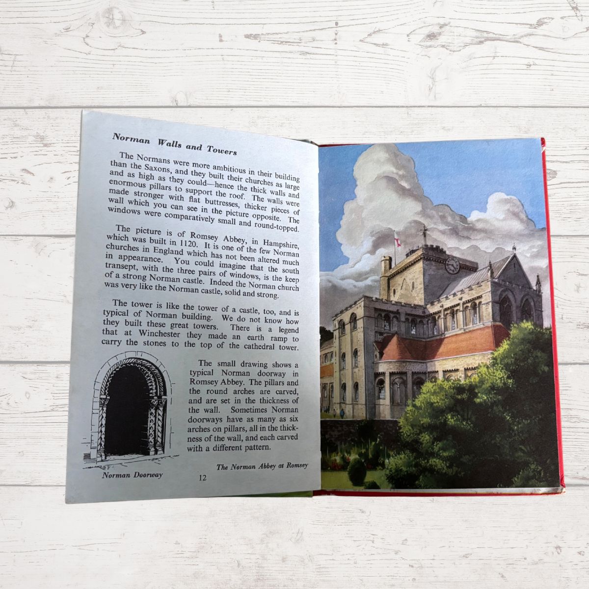 An open vintage illustrated book spread showing text about Norman walls and towers, with a large color illustration of a Norman castle and garden on the right page, photographed on a white wood surface.