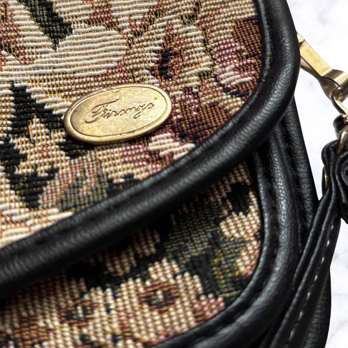 Close-up of a tapestry bag with a floral pattern in beige, brown, and black, featuring black leather trim and a gold-tone oval logo plate.