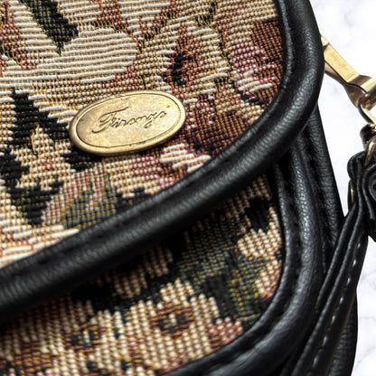 Close-up of a tapestry bag with a floral pattern in beige, brown, and black, featuring black leather trim and a gold-tone oval logo plate.