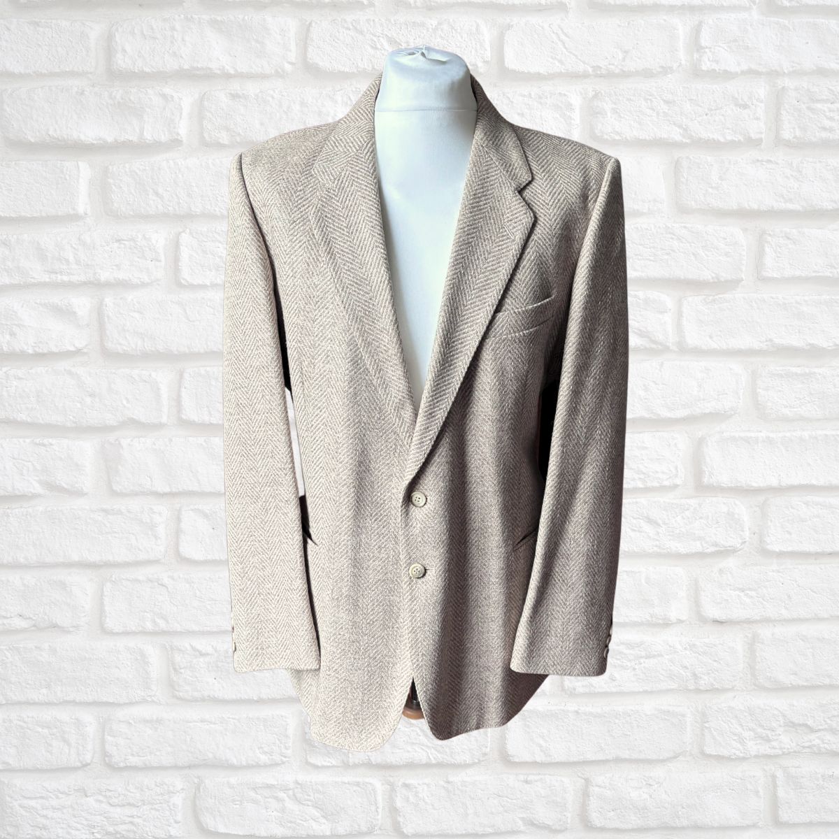 A light beige textured herringbone blazer displayed on a white mannequin torso against a white brick wall background.