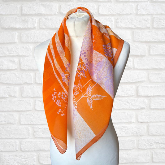 Orange scarf with floral patterns on a mannequin against a white brick wall.