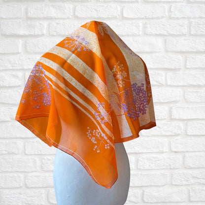 Orange scarf with floral patterns draped over a mannequin head against a white brick wall.
