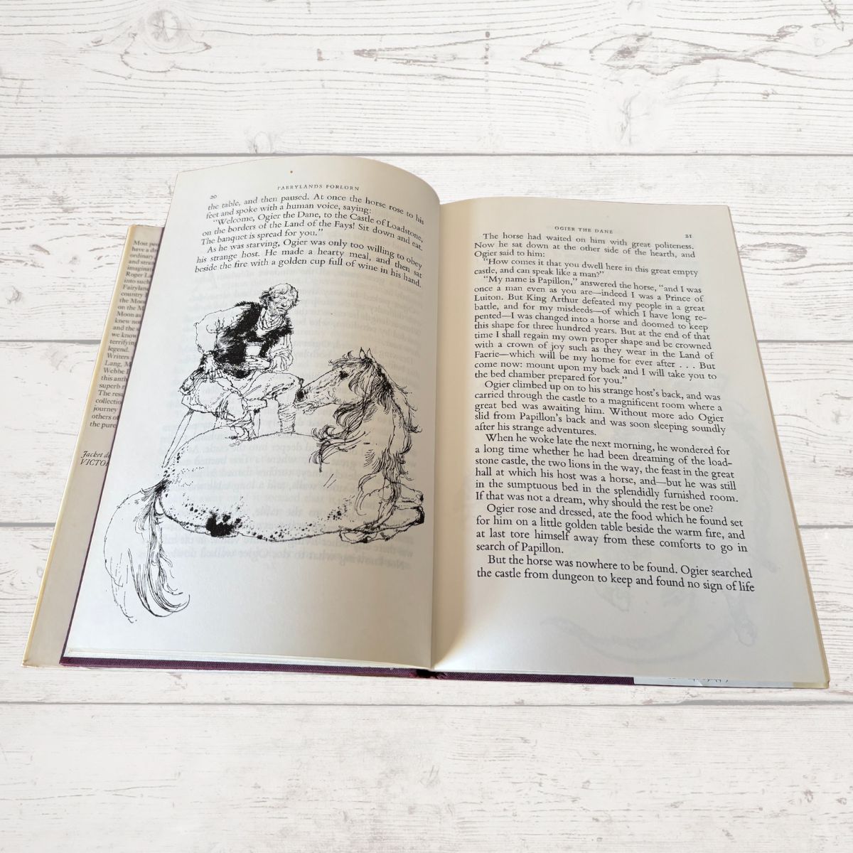 An open vintage book showing printed text and a black ink illustration of two figures, photographed on a light whitewashed wooden surface.