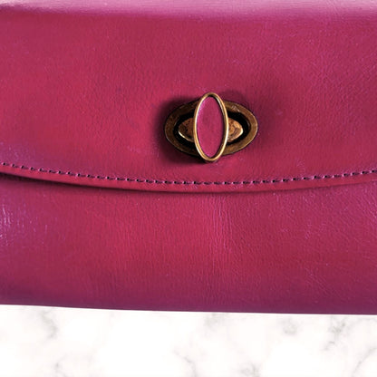 A close-up of a red leather clutch or wallet featuring a distinctive gold-tone metal turn-lock clasp on the front flap, set against a white marble surface.
