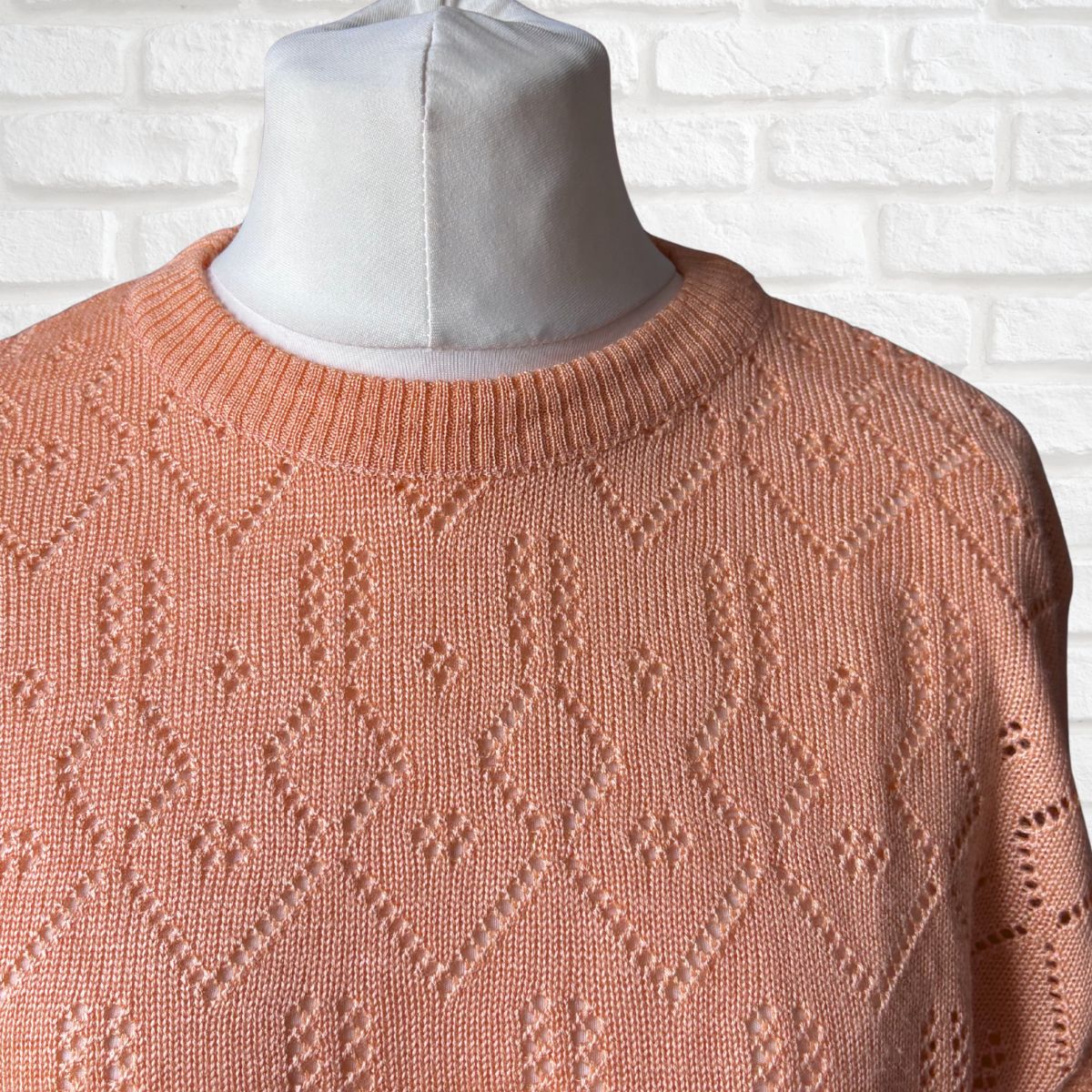 A peach-colored knit sweater with a textured geometric diamond pattern, displayed on a white mannequin torso against a white brick wall background.