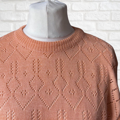 A peach-colored knit sweater with a textured geometric diamond pattern, displayed on a white mannequin torso against a white brick wall background.