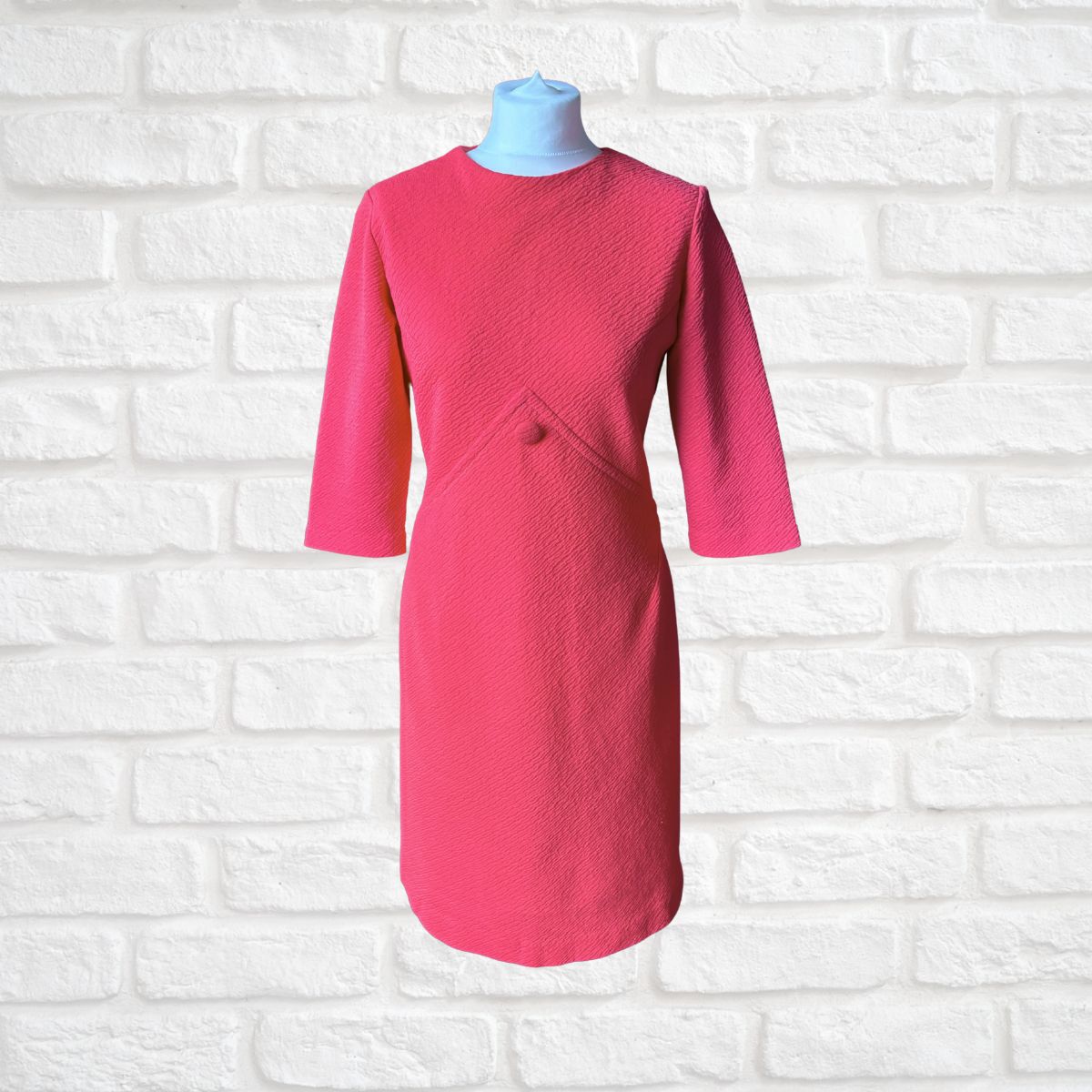 A bright pink textured dress with three-quarter sleeves and a small button detail at the waist, displayed on a light blue mannequin torso against a white brick wall background.