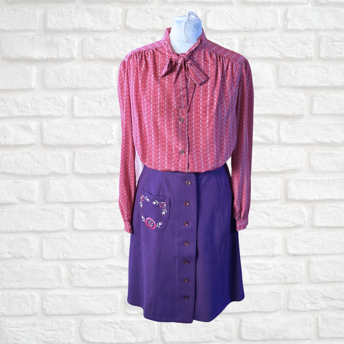 Vintage outfit with a pink patterned blouse and purple skirt on a white brick wall background
