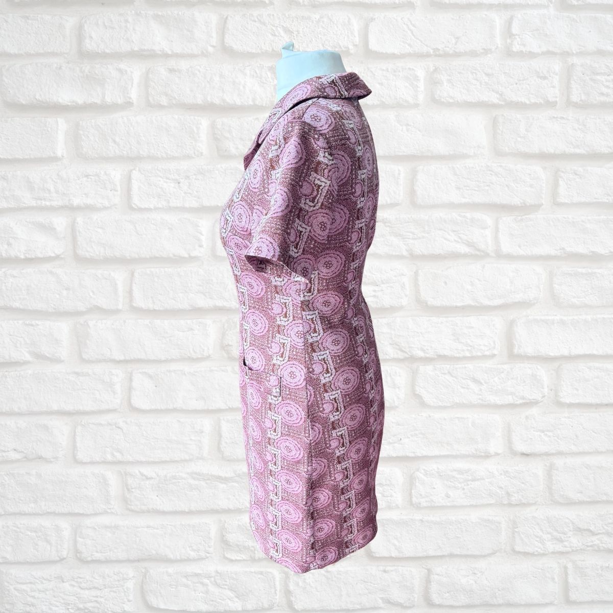 A short-sleeve collared dress  in a pink and purple paisley pattern, displayed on a mannequin torso against a white brick wall background.