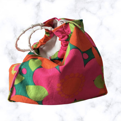 A retro-style fabric bag  with a bold mid-century floral pattern in pink, orange, green, and yellow, featuring a white bamboo-look handle, set against a white marble background.