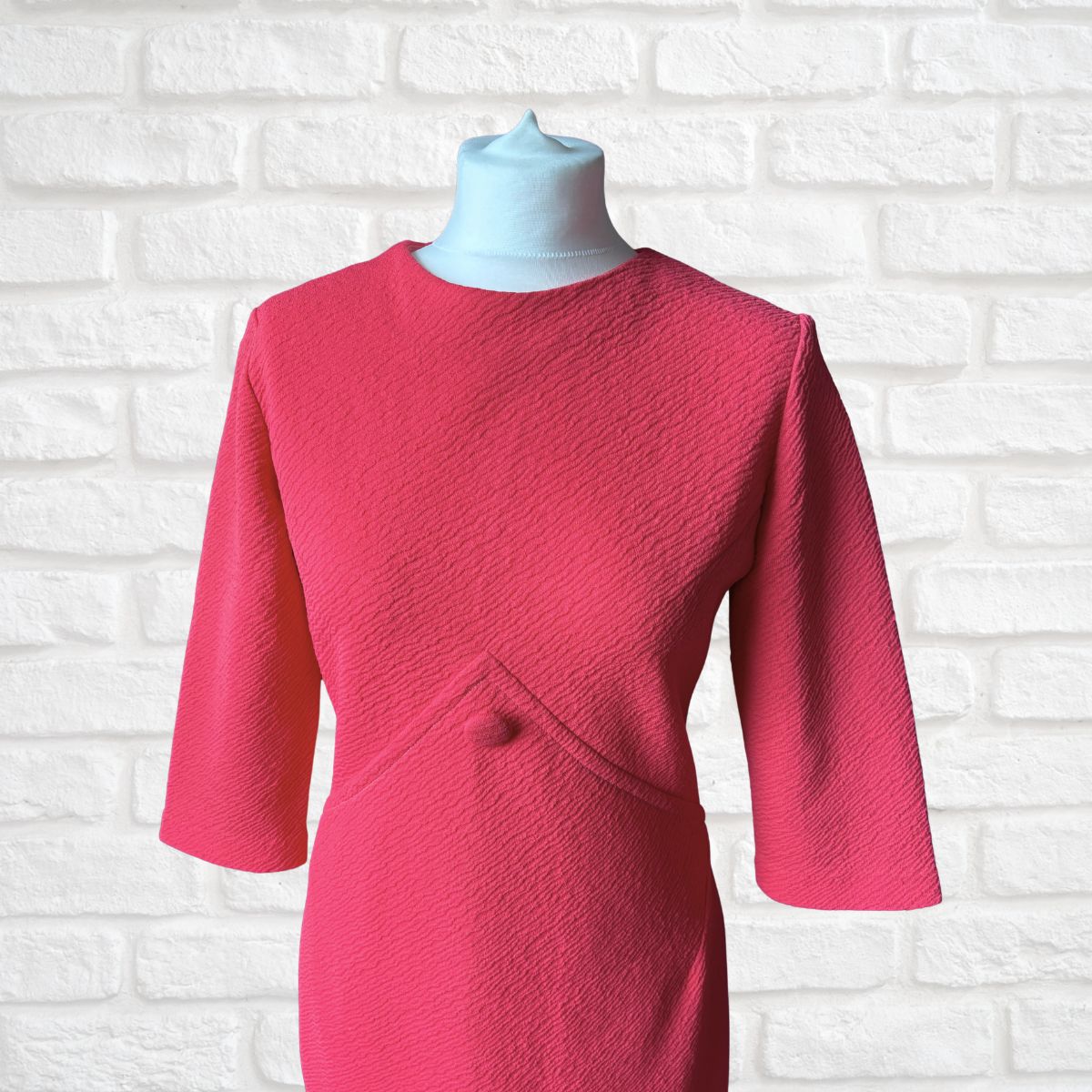 A bright pink textured dress with three-quarter sleeves, displayed on a light blue mannequin torso against a white brick wall background.