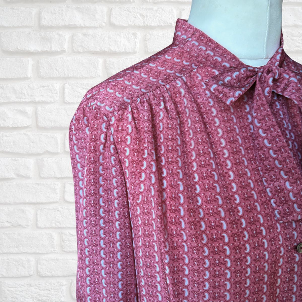 Close-up of a pink and grey patterned blouse on a mannequin against a white brick wall.