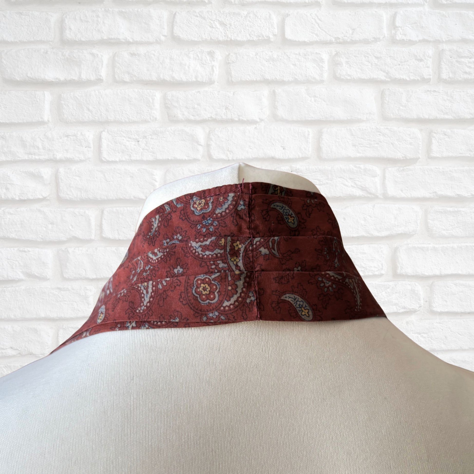 The back of vintage silk cravat featuring a paisley print in red, grey, yellow, and blue, displayed on a mannequin against a white brick wall background.