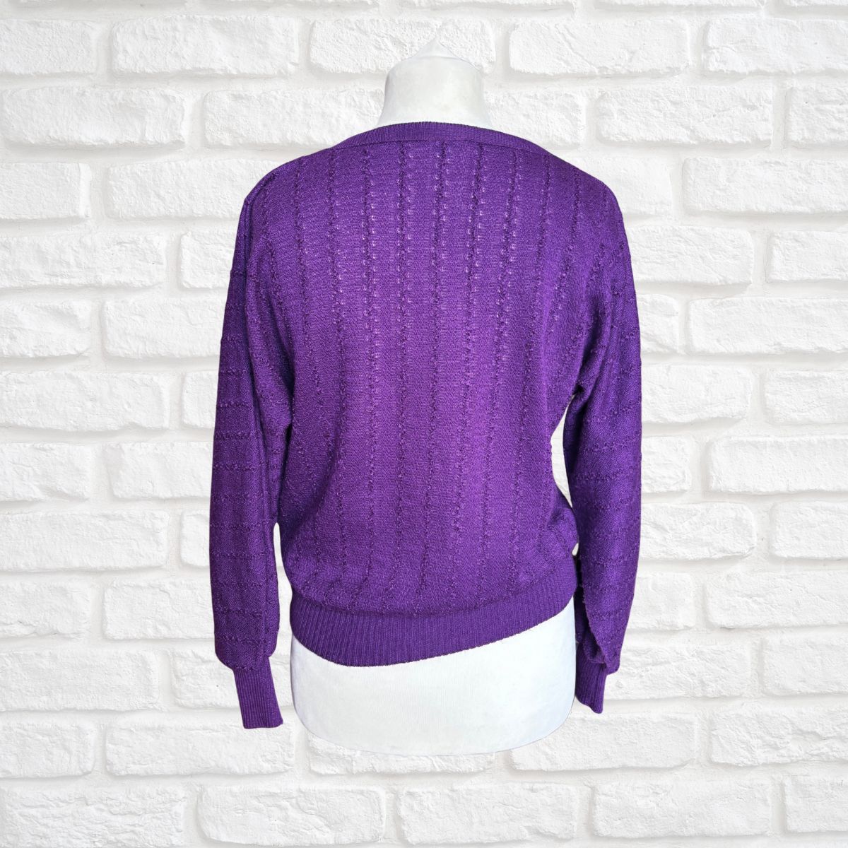 A purple cable-knit sweater displayed on a white mannequin torso against a white brick wall background.