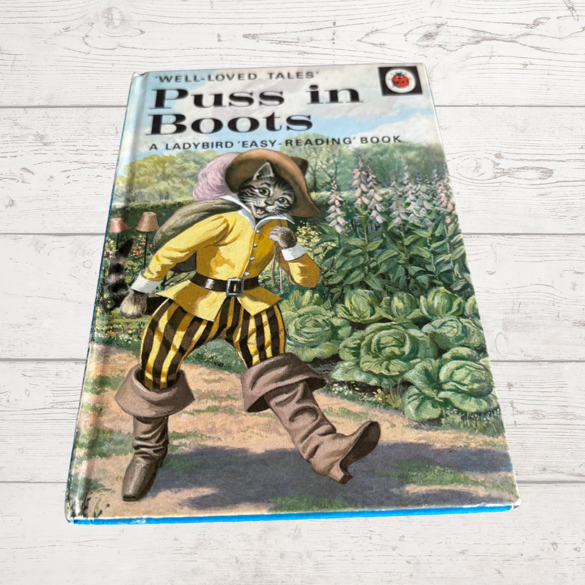 A vintage children's book titled 'Puss in Boots' from the Well-Loved Tales series, featuring an illustration of a cat dressed in a yellow and black striped outfit in a garden, photographed on a white wood plank surface.