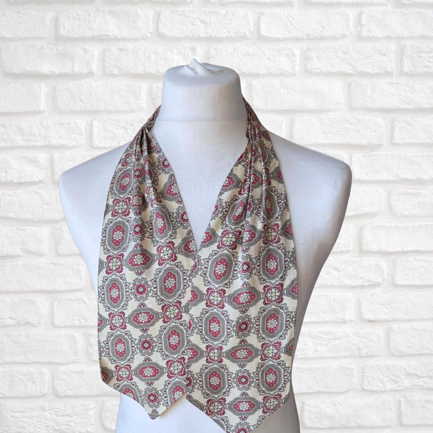 A vintage 60s mod style cream cravat with a paisley print featuring baroque style red, grey, and green patterns, displayed on a mannequin against a white brick wall background.