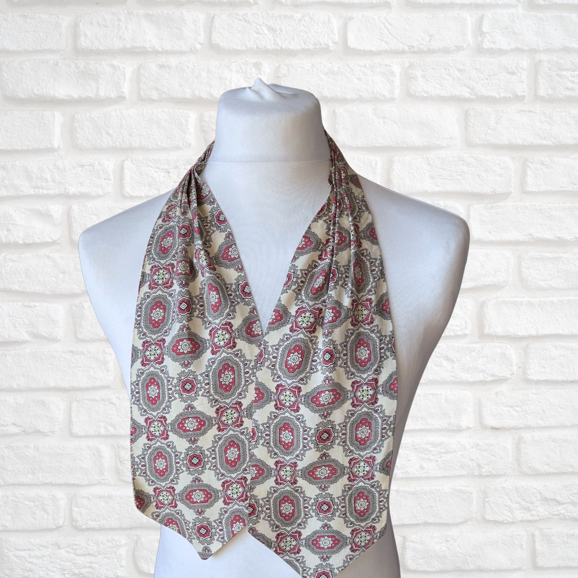 A vintage 60s mod style cream cravat with a paisley print featuring baroque style red, grey, and green patterns, displayed on a mannequin against a white brick wall background.