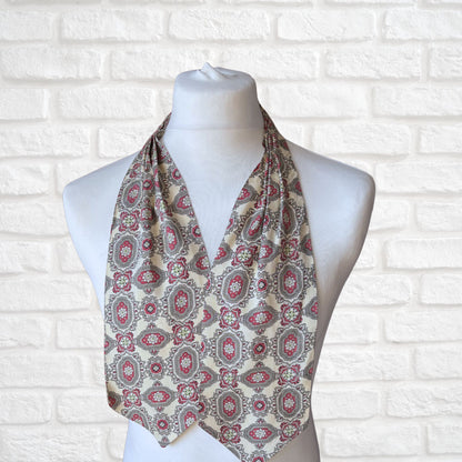 A vintage 60s mod style cream cravat with a paisley print featuring baroque style red, grey, and green patterns, displayed on a mannequin against a white brick wall background.