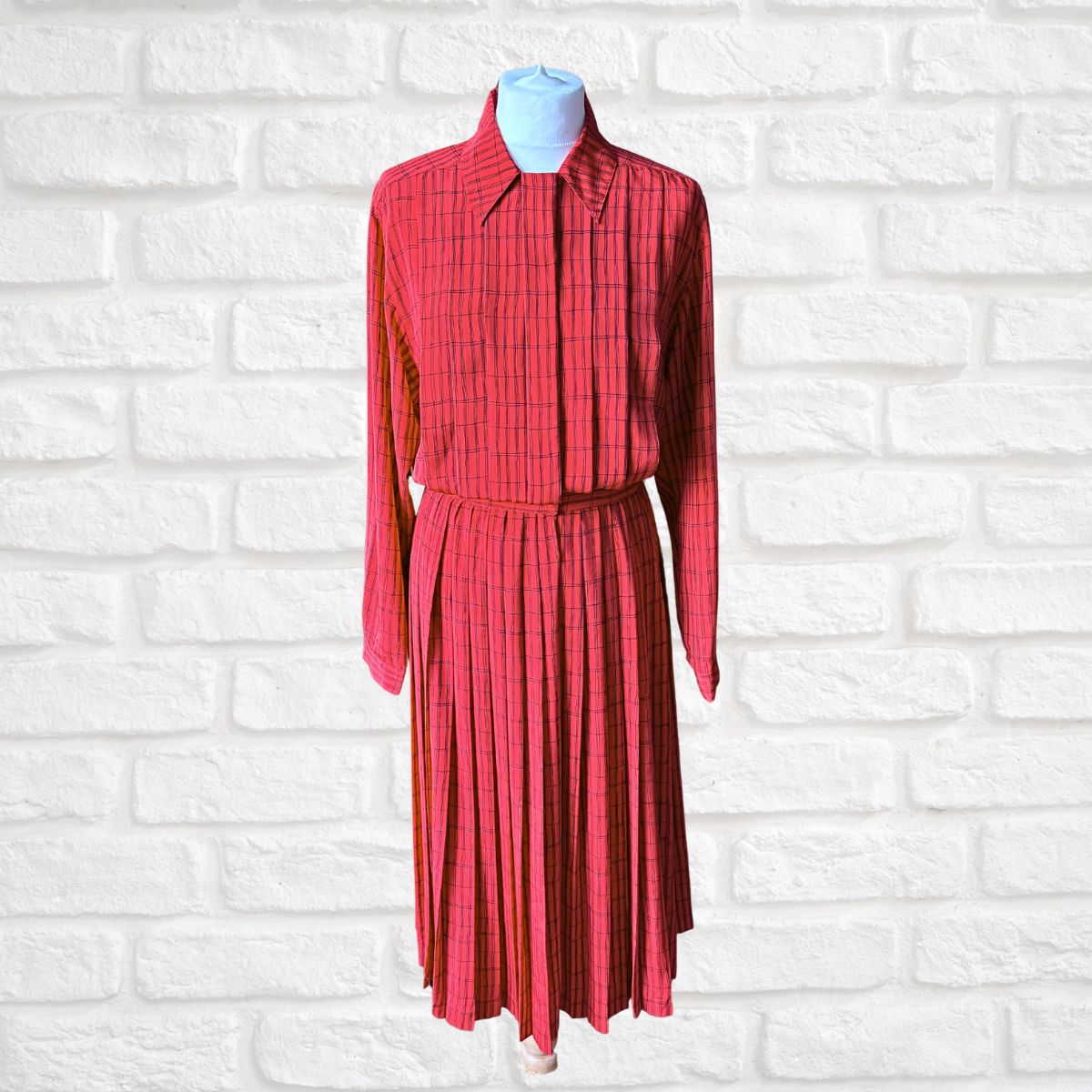 A red and black plaid long-sleeve collared shirt and matching pleated skirt set displayed on a mannequin torso against a white brick wall background.