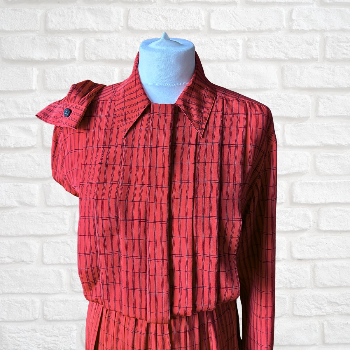 A red and black plaid long-sleeve dress displayed on a light blue mannequin torso against a white painted brick wall.