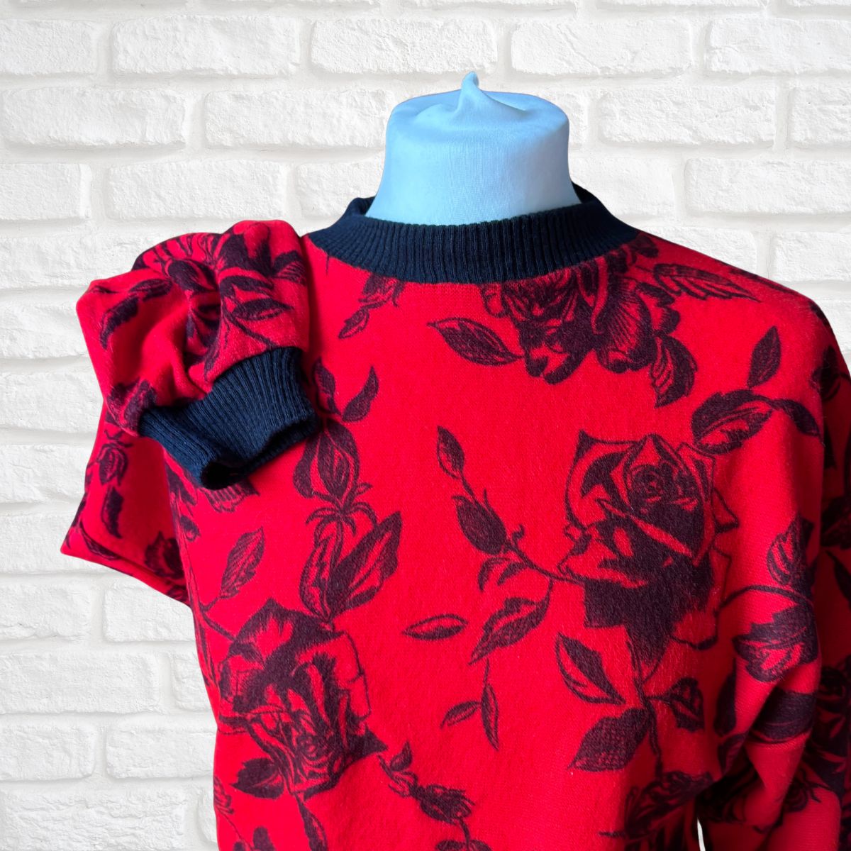 A red knit sweater with a black rose and leaf pattern, featuring a black ribbed crew neck and a matching black ruffled sleeve detail, displayed on a light blue mannequin torso against a white brick wall background.