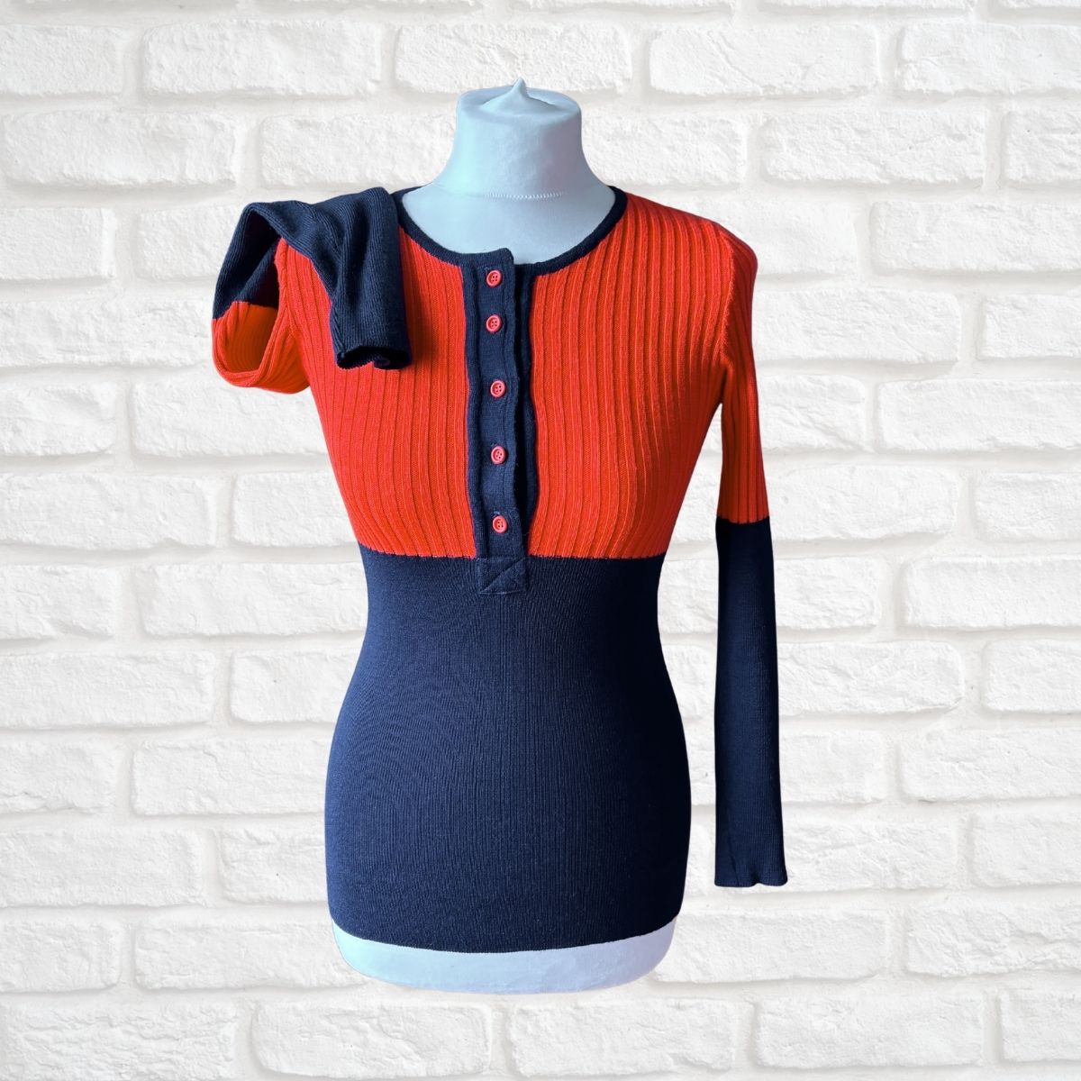 A color-block ribbed knit sweater displayed on a mannequin torso against a white brick wall. The top portion is bright orange-red with a three-button henley neckline, while the lower portion and sleeves are dark navy blue.