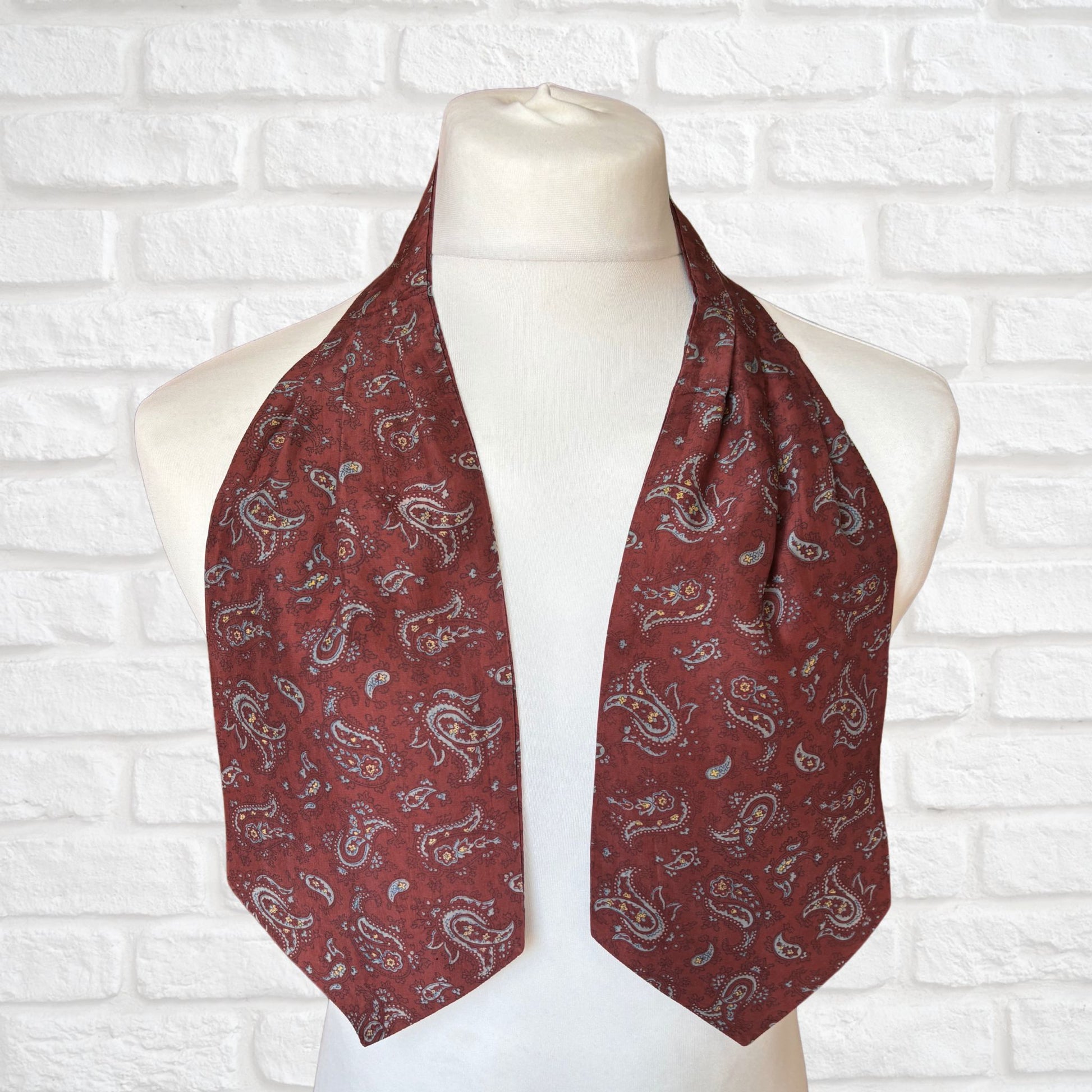 A vintage silk cravat featuring a paisley print in red, grey, yellow, and blue, displayed on a mannequin against a white brick wall background.