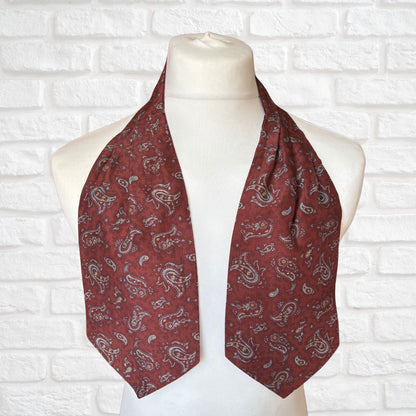 A vintage silk cravat featuring a paisley print in red, grey, yellow, and blue, displayed on a mannequin against a white brick wall background.