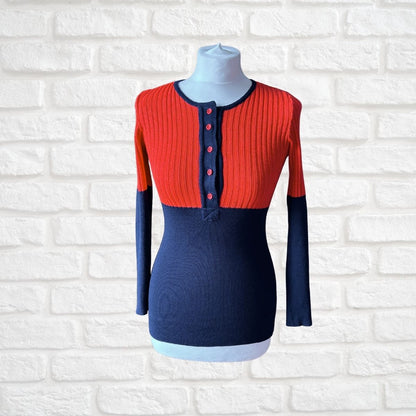 A color-blocked ribbed knit long-sleeve top displayed on a dress form mannequin against a white brick wall background. The top features a bright red yoke with a three-button placket, navy blue sleeves and lower body, and a light blue inner hem.