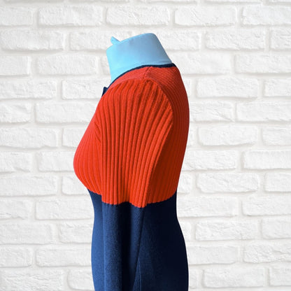 A colorblock ribbed knit sweater displayed on a mannequin torso against a white brick wall background. The top portion is a vibrant orange-red, the bottom portion is navy blue, and the collar is light blue.