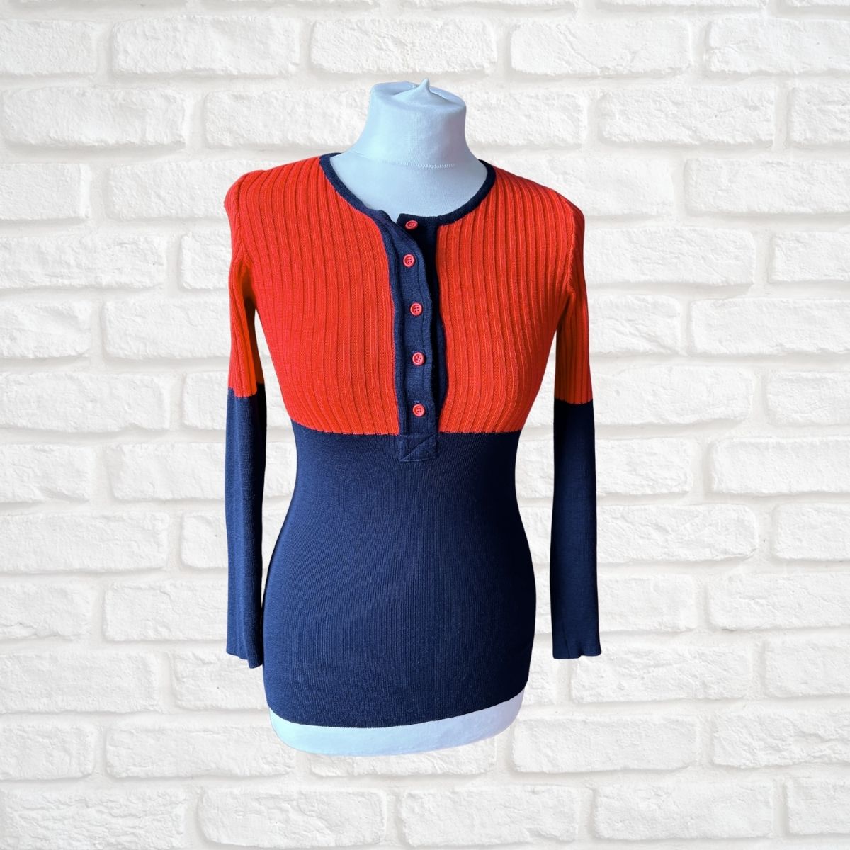 A colorblock ribbed knit long-sleeve top displayed on a white mannequin torso against a white brick wall. The top features a vibrant orange-red yoke with a buttoned henley neckline, contrasting with a navy blue body and matching navy sleeves.