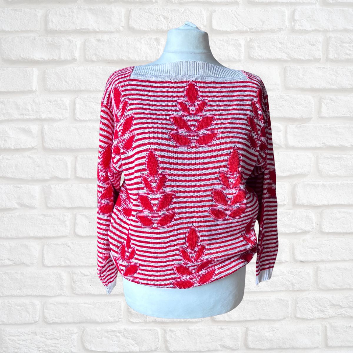 A red and white striped knit sweater with red floral motifs, displayed on a white mannequin torso against a white brick wall background.