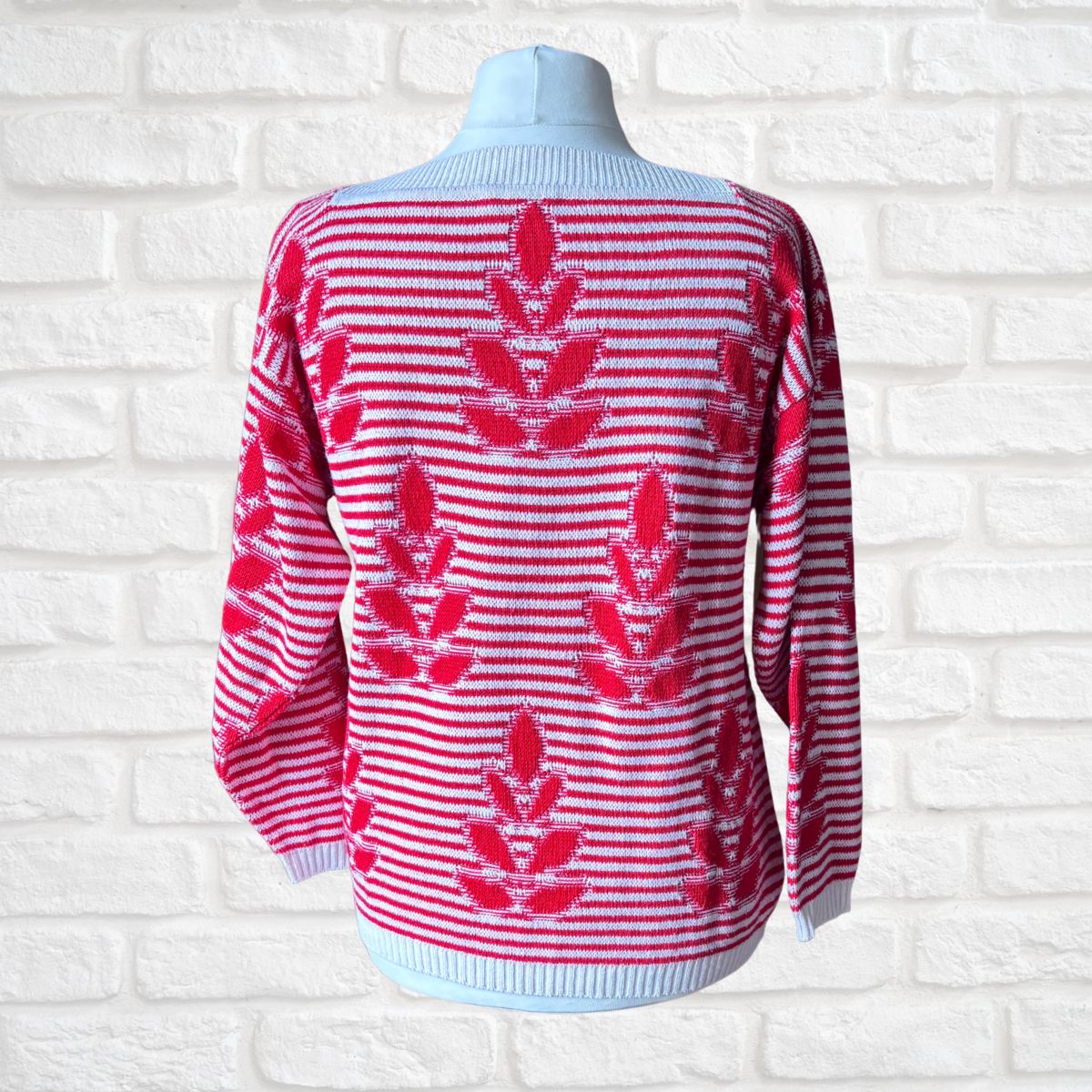 A red and white striped knit sweater with red leaf motifs, displayed on a light blue mannequin torso against a white brick wall background.