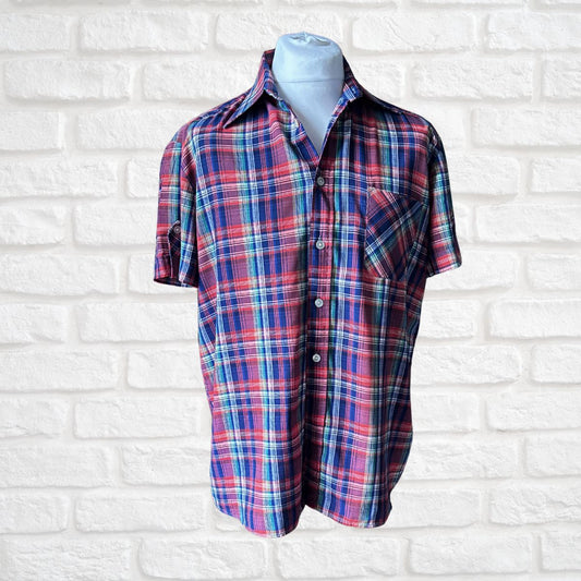 A short-sleeve button-up plaid shirt in red, blue, and white, displayed on a light blue mannequin torso against a white brick wall background.