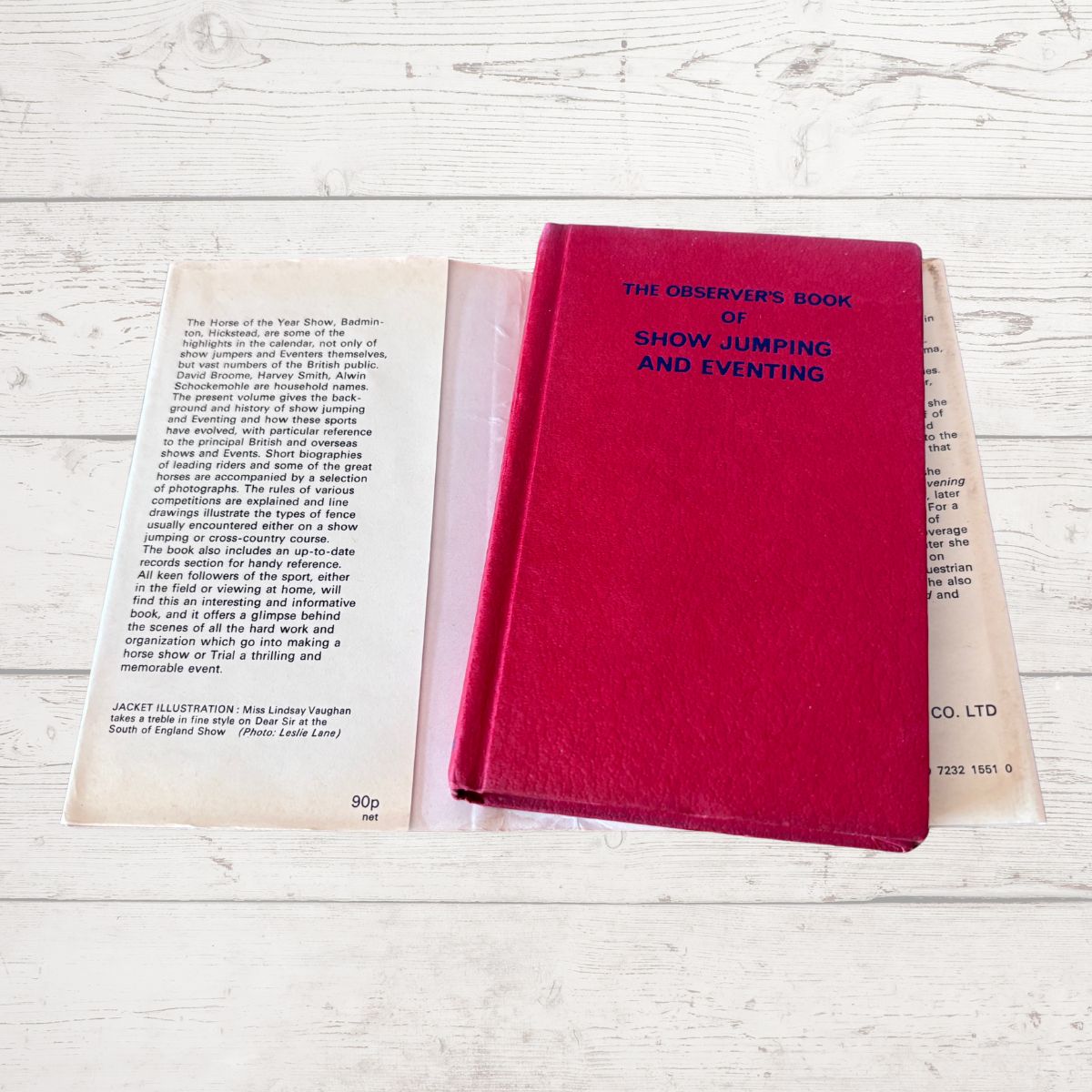 A vintage hardcover book titled 'The Observer's Book of Show Jumping and Eventing' in a deep magenta-red cloth cover, placed on top of its open printed pages on a whitewashed wooden surface.