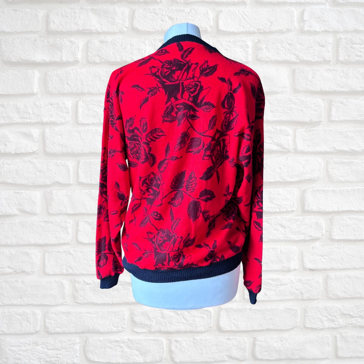 A red long-sleeve sweatshirt  with a black rose pattern, displayed on a light blue mannequin torso against a white brick wall background.
