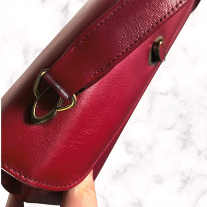 A close-up of a red leather handbag held by a hand against a white marble background, showing a brass D-ring and strap attachment.