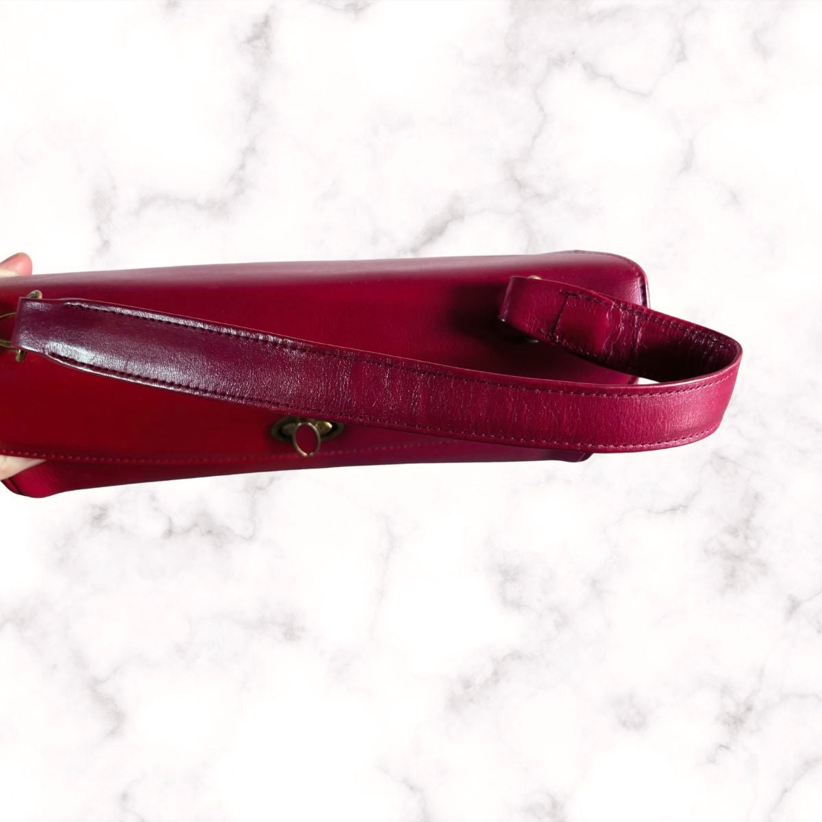 A close-up of a deep red  leather handbag strap with a small gold heart-shaped hardware accent, held against a white marble background.