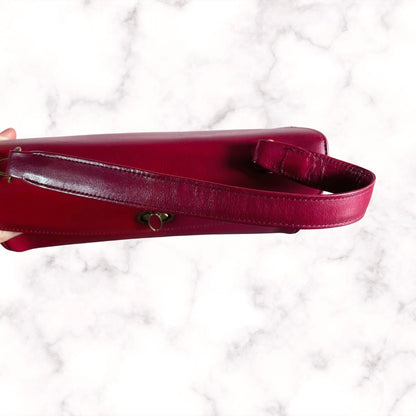A close-up of a deep red  leather handbag strap with a small gold heart-shaped hardware accent, held against a white marble background.