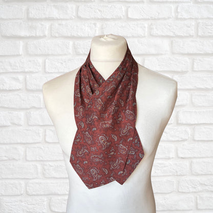 A vintage silk cravat featuring a paisley print in red, grey, yellow, and blue, displayed on a mannequin against a white brick wall background.