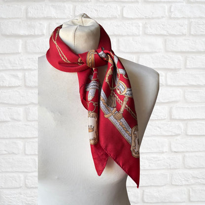 Red scarf with gold and white patterns on a mannequin against a white brick wall.