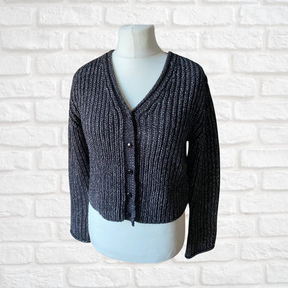 A 90s black cropped long-sleeved vintage cardigan with ribbed pattern and silver metallic thread, displayed on a mannequin against a white brick wall background.