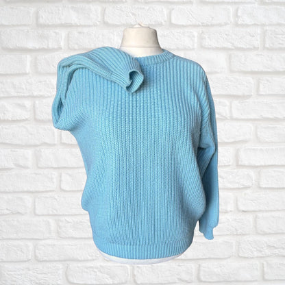 A light blue ribbed knit sweater displayed on a white mannequin torso against a white painted brick wall background.