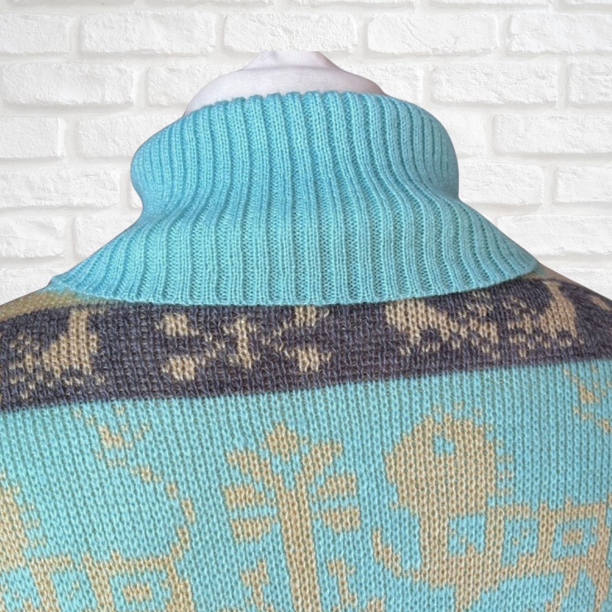 A close-up of a knit turtleneck sweater on a mannequin, featuring a light blue ribbed neck and a patterned body with beige motifs and a dark gray horizontal band.